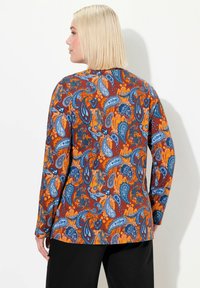 Langärmliges Oberteil mit einem Paisleymuster in Orange, Blau und Braun auf einem dunkleren Hintergrund. Hergestellt aus strukturiertem Stoff.
