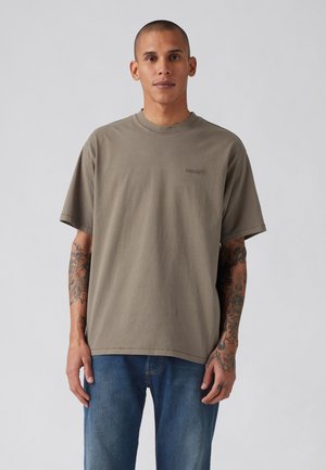 Mand med tatoveringer iført en enkel, overdimensioneret brun t-shirt og blå jeans, der står mod en ensfarvet lys baggrund.