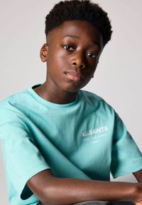 Ljus tealfärgad bomullströja med korta ärmar och rundad halsringning. Framsidan har texten "ALLSAINTS" i vitt med ytterligare varumärkesinformation.