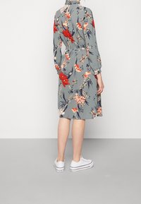 Vestido floral en gris con flores rojas y rosas, de mangas largas, cintura elástica y largo hasta la rodilla. Combinado con zapatillas blancas.