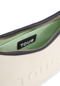 Bolso sintético color crema con logo "TOUS" en relieve, cremallera dorada y ribete negro; el interior presenta un forro verde.