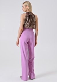 Femme avec de longs cheveux blonds, portant un haut sans manches en velours marron et un pantalon large taille haute lilas, se tenant devant un fond uni.