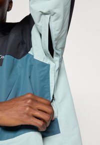 Kurtka wodoodporna w kolorze turkusowo-czarnym, z zamkniętą kieszenią na zamek błyskawiczny i teksturowanym materiałem.