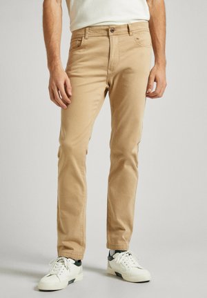 Pantalones de mezcla de algodón color caqui con corte slim, que cuentan con cremallera, diseño de cinco bolsillos y un sutil detalle de costura, combinados con zapatillas blancas.