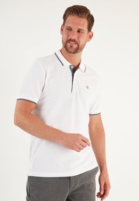 Bílá polo košile se šedým límcem a lemy, krátkými rukávy a třemi knoflíky u krku. Obsahuje malé logo na hrudi.