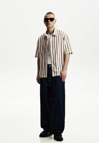 Short-sleeve, brown and white striped button-up shirt over a white t-shirt, paired with wide-leg dark denim pants and black shoes.