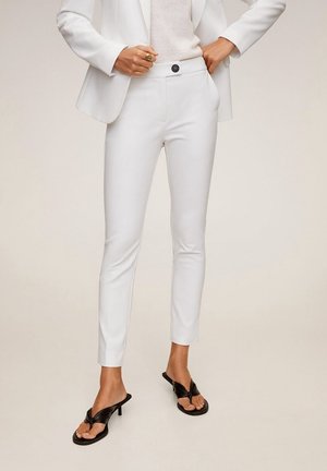 Woman wearing white tailored pants, white blazer, and black heeled sandals, standing with one hand in pocket and one hand on waist.