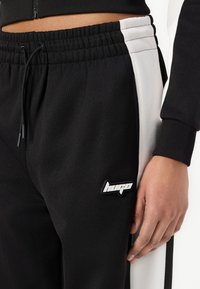 Crne sportske hlače s elastičnom trakom u struku, bijelim bočnim trakama i gumenim "Hugo" logom na lijevom bedru. Glatka, teksturirana tkanina.