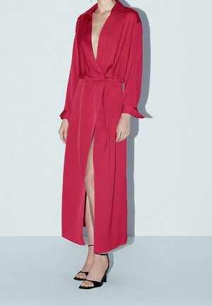 Femme porte une longue robe portefeuille rouge avec un décolleté en V profond et une fente latérale, associée à des talons ouverts noirs sur un fond clair uni.