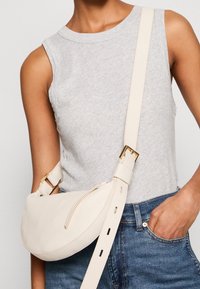 Bolso cruzado de cuero color crema con forma curva, un bolsillo frontal con cremallera y tira ajustable con herrajes dorados.