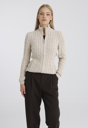 Jeune femme aux longs cheveux blonds portant un pull zippé beige en maille torsadée et un pantalon ample foncé, debout devant un fond uni.