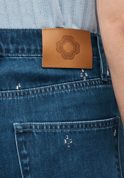 Κοντινό πλάνο μπλε τζιν με λεπτομέρειες από ασημένιες καρφίτσες και καφέ δερμάτινο ταμπελάκι με λουλουδένιο λογότυπο στη μέση.