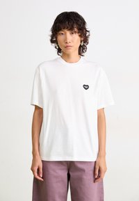 T-shirt en coton blanc à manches courtes, avec un petit logo en forme de cœur noir sur la poitrine gauche. Design simple et décontracté.
