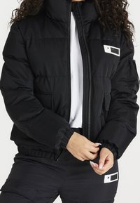 Svart vadderad jacka med hög krage, dragkedjestängning, elastiska ärmslut och sidofickor. Har en vit etikettdetalj på bröstet.