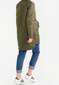 Manteau long vert olive avec une texture lisse, boutons sur le devant et poches latérales, associé à un jean bleu retroussé et des baskets noir et blanc.