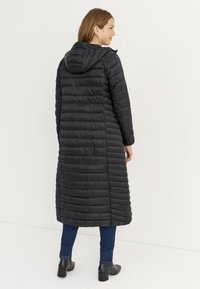 Sort puffer-jakke med hætte, der har et langt, lige design og vandret quiltet mønster. Båret over blå jeans og sorte ankelstøvler.