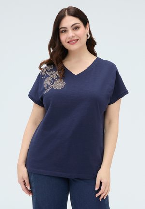 Kobieta uśmiechająca się, ubrana w granatową koszulkę z dekoltem w serek i złotym kwiatowym haftem na ramieniu oraz niebieskie dżinsy na jasnym tle.