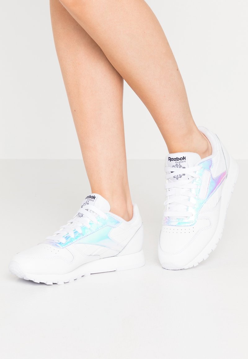 Jambes portant des baskets blanches Reebok Classic avec des détails latéraux brillants et irisés, debout sur un sol gris clair sur fond blanc.