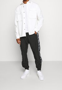 Man in een wit overhemd met knopen, zwarte sportbroeken met witte logo-stripes en witte sneakers, met de handen in de zakken.