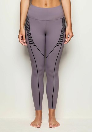 Frau, die barfuß steht und hochgeschnittene mauvefarbene Leggings mit schwarzen geometrischen Linien und gestreiften Akzenten an den Seiten trägt.