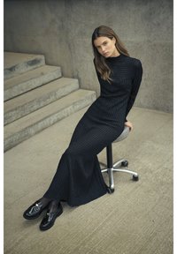 Schwarzes, strukturiertes Maxikleid mit langen Ärmeln und figurbetonter Silhouette. Dazu glänzende schwarze Loafers auf einem silbernen Rollstuhl.