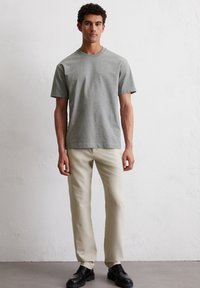 Szary bawełniany T-shirt z krótkim rękawem i okrągłym dekoltem, zestawiony z jasnobeżowymi spodniami i czarnymi, sznurowanymi butami na neutralnym tle.