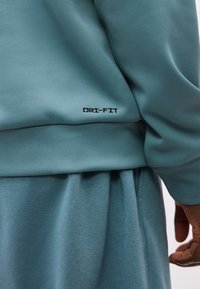 Κοντινή λήψη σε αθλητική ενδυμασία σε τυρκουάζ με μαύρο λογότυπο "DRI-FIT" στο φούτερ, που δείχνει την υφή του υφάσματος και μέρος του χεριού ενός ατόμου.