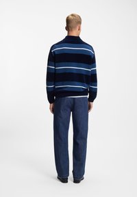 Pull en maille à rayures marines avec des rayures horizontales bleu clair et blanches. Porté avec un pantalon en denim bleu. Fond uni.