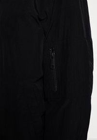 Veste noire en tissu texturé et léger. Le design comprend une poche zippée sur le bras gauche et une poche latérale cachée.