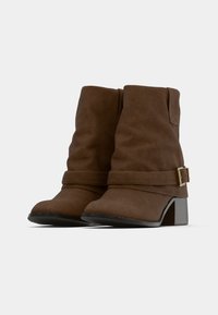 Brūnas zamša zābaki līdz potītei ar salocītu dizainu, sānu siksnu un zelta tonīša sprādzi kā akcentu, kā arī ar masīvu melnu papēdi.