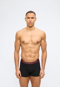 Μυώδης νέος με κοντά μαλλιά που φοράει μαύρα μποξεράκια Tommy Hilfiger, στέκεται μπροστά σε απλό ανοιχτό φόντο.