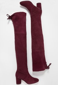 Botas de ante burdeos por encima de la rodilla, con tacón cuadrado y un lazo decorativo en la parte superior, de textura suave en todo su diseño.