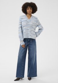 Femme aux cheveux bouclés portant un pull à col en V rayé bleu et gris, un jean à jambes larges et des talons aiguilles noirs, se tenant avec une main sur la hanche.