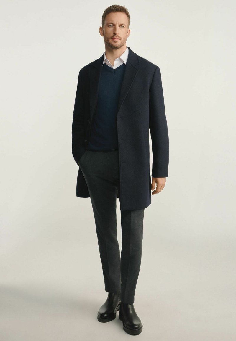 Granatowy wełniany płaszcz na granatowym swetrze i białej koszuli, zestawiony z ciemnoszarymi spodniami i czarnymi botkami. Prosty, dopasowany fason.