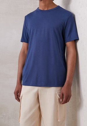 Hombre vestido con una camiseta azul lisa de manga corta y pantalones cargo beige, de pie contra una pared de color claro.