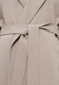 Κοντινό πλάνο μπεζ υφασμάτινης ζώνης δεμένης γύρω από ένα παλτό με διαγώνια υφή ύφανσης και επικαλυπτόμενες πέτα.