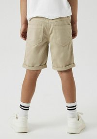 Shorts kaki avec des ourlets retroussés, deux poches arrière et une texture lisse, associés à des baskets blanches et des chaussettes rayées.