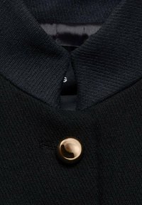 Černý pletený límec oděvu s hladkým saténovým podšívkou a zlatým knoflíkem na zapínání. Textura je žebrovaná a matná.