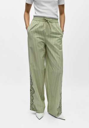 Nő világoszöld tűcsíkos, széles szárú nadrágban, oldalt csipkedíszítéssel, fehér hegyes orrú cipővel és fehér rövid ujjú felsővel.