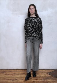 Zwart-crèmekleurige trui met zebrapatroon, grijze spijkerbroek en zwarte enkellaarzen. Model staat tegen een witte muur met textuur.