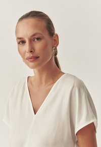 Weißes V-Ausschnitt-Bluse mit kurzen Ärmeln, glatte Textur; kombiniert mit großen, goldfarbenen, strukturierten ovalen Ohrringen. Einfaches und elegantes Design.