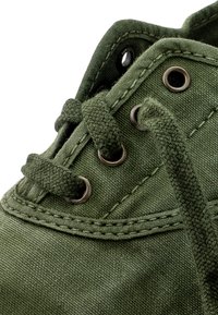 Primer plano de una zapatilla de tela verde oliva con cordones gruesos tejidos pasando por ojales de metal y costuras detalladas.