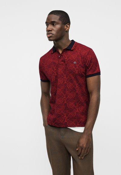 Άνδρας που φοράει κόκκινη μπλούζα πόλο με σχέδια, σκούρο γιακά και μανσέτες, καφέ παντελόνια, στέκεται με τα χέρια στις τσέπες, κοιτώντας προς τα αριστερά.