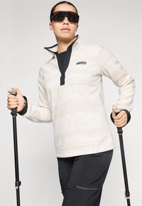 Witte fleece pullover met tribale patroon, zwarte kraag en sluiting, gecombineerd met zwarte broek. Model houdt trekkingstokken vast en draagt een zonnebril.