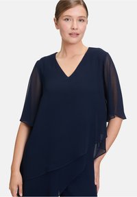 Granatowa bluzka z dekoltem w kształcie litery V, krótkimi falbankowymi rękawami i warstwowym wzorem. Wykonana z lekkiego materiału o miękkiej fakturze.