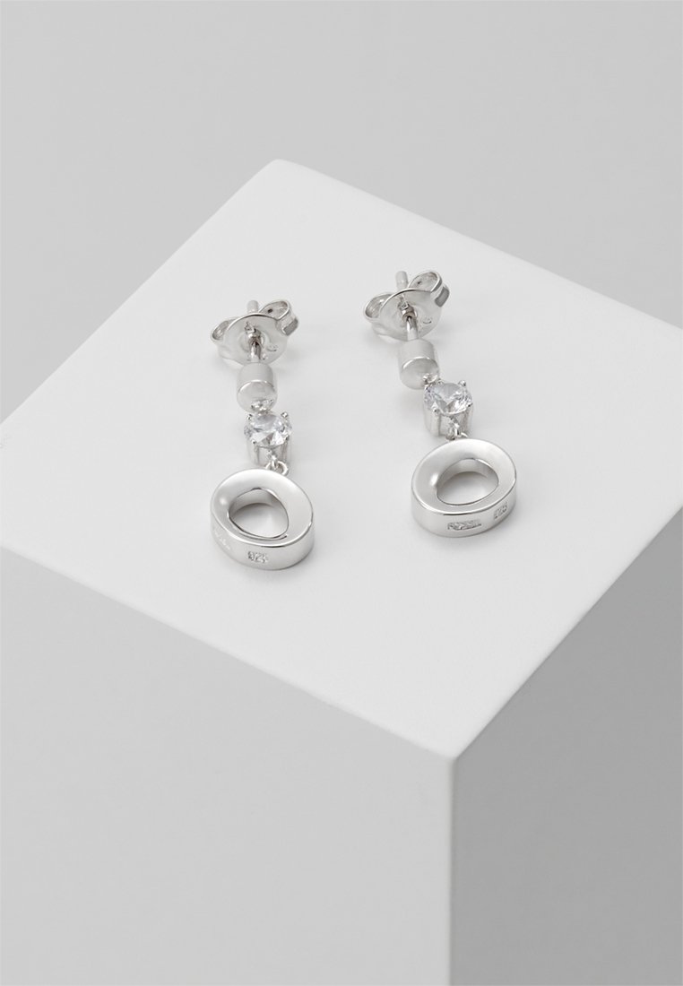 Boucles d'oreilles clous en argent avec petites pierres précieuses rondes et transparentes et pendentifs creux circulaires, exposées sur une surface cubique blanche.