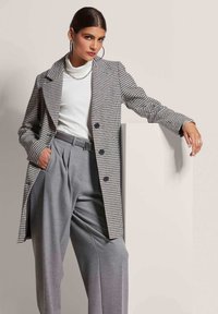 Zwart-wit houndstooth-patroon trenchcoat met notch-kraag, gecombineerd met hoogsluitende grijs geplooide broek en een witte geribbelde turtleneck.