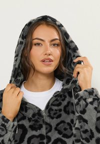 Hoodie gris à motif léopard en tissu doux et texturé, avec une fermeture éclair et une capuche à cordon, porté par-dessus une chemise blanche.