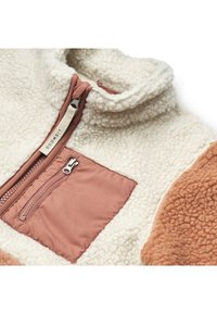 Pulover din fleece pufos, crem, cu guler înalt, buzunar contrastant terracotta și un buzunar cu fermoar pe piept. Detaliile texturate ale suprafeței sunt vizibile.