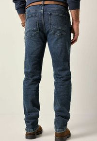 Vaqueros de mezclilla en azul oscuro, corte ajustado. Presenta un diseño de cinco bolsillos, costuras sutiles y una textura ligeramente descolorida. Combinados con botas marrones.